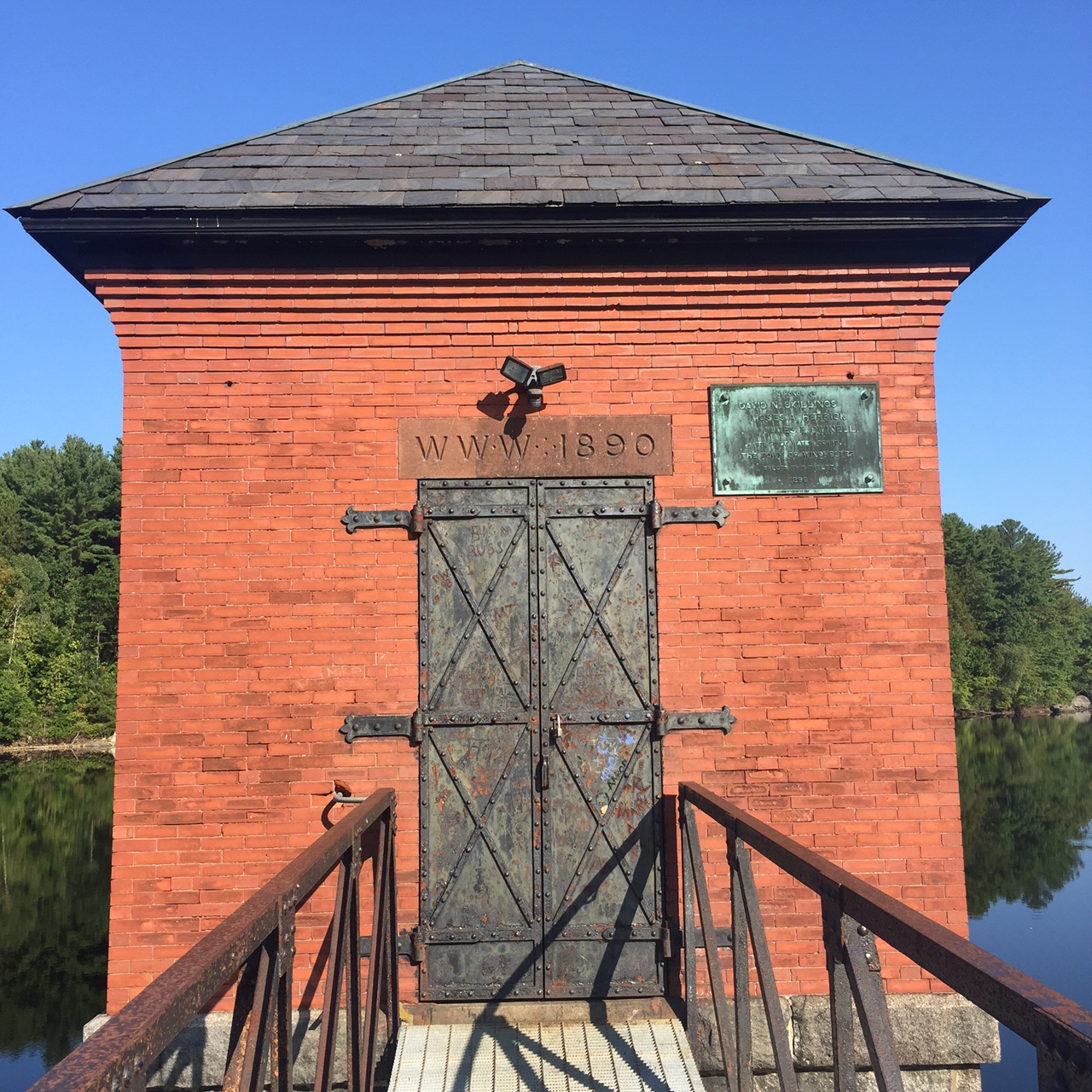 Small brick building surrounded by water, with a carved plaque reading 'WWW 1890' over a metal door. ()